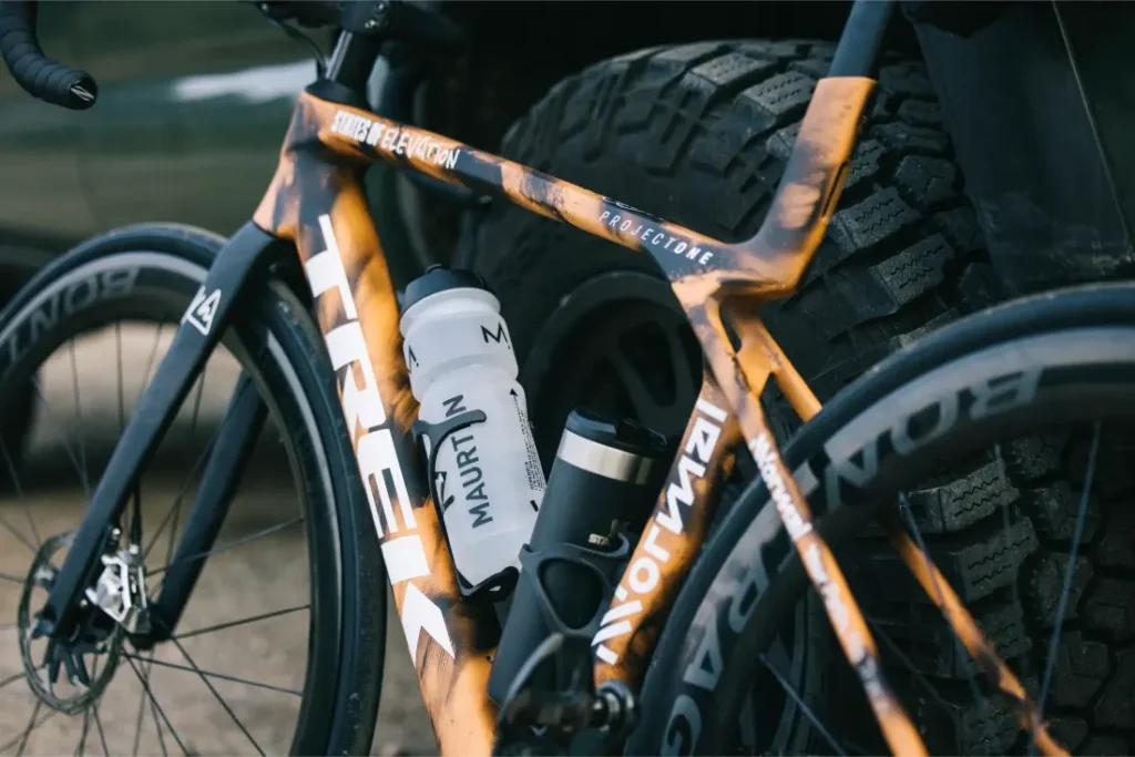 Un primer plano de un cuadro de bicicleta de carretera TREK naranja y negro con dos bidones de agua montados, colocado junto a un neumático de vehículo, evoca el espíritu de States of Elevation - Kilian Jornet.