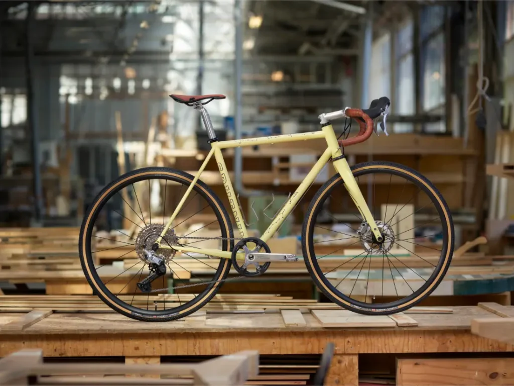 Una bicicleta de carretera amarilla con manillar en caída, con grupos de carretera Shimano CUES, se exhibe en el interior sobre un banco de trabajo de madera en el entorno de un taller.