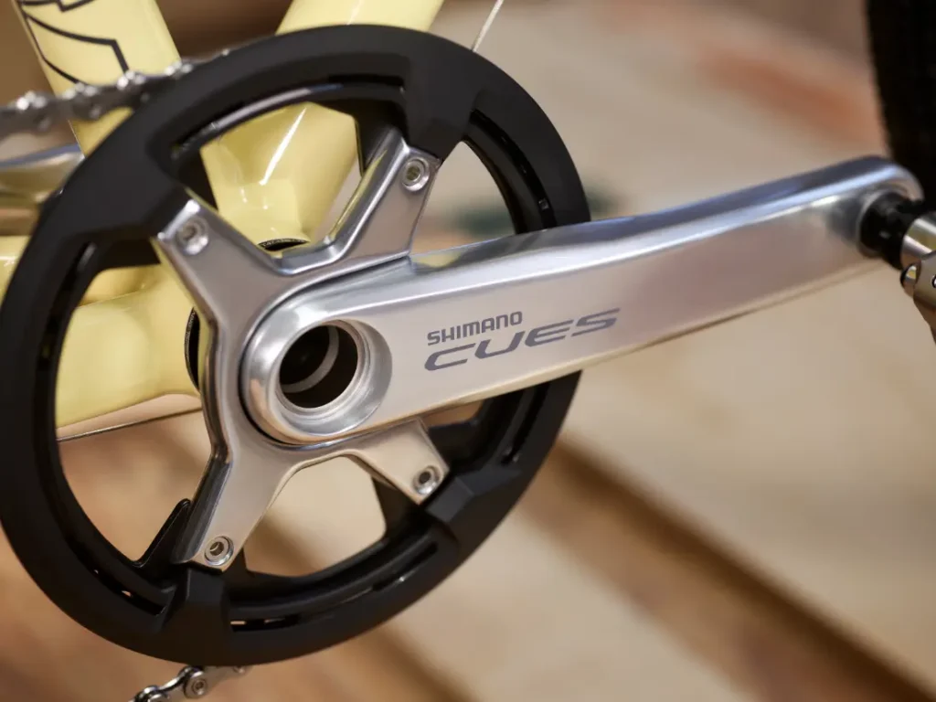 Primer plano de un plato de bicicleta Shimano CUES con brazo de pedal acoplado, fabricado en Aluminio Pulido y montado en un cuadro de bicicleta amarillo claro, perfecto para quienes actualizan sus grupos de carretera.