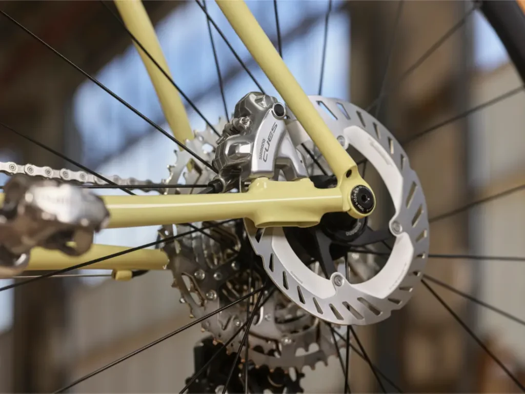 Primer plano de la rueda trasera de una bicicleta que muestra el rotor del freno de disco, la pinza, parte del cassette del cambio y componentes Shimano CUES de grupos de carretera.