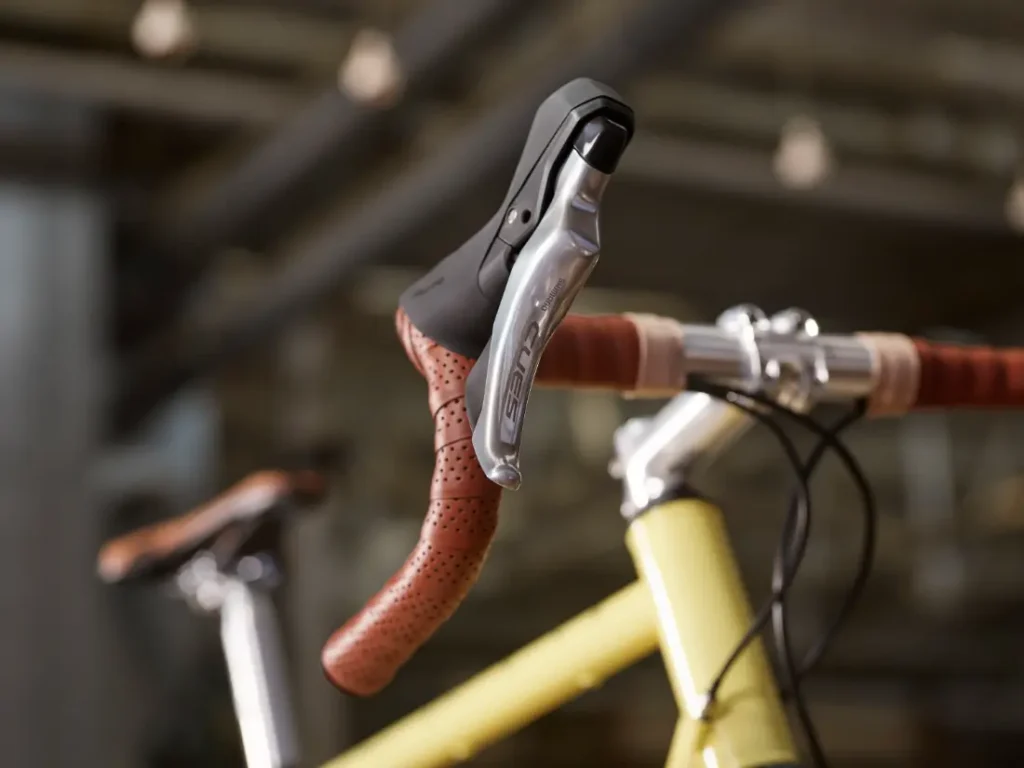 Primer plano de la cinta de manillar marrón de una bicicleta de carretera, la palanca de freno plateada y parte de un cuadro amarillo con grupos de carretera Shimano CUES, sobre un fondo borroso de interior.
