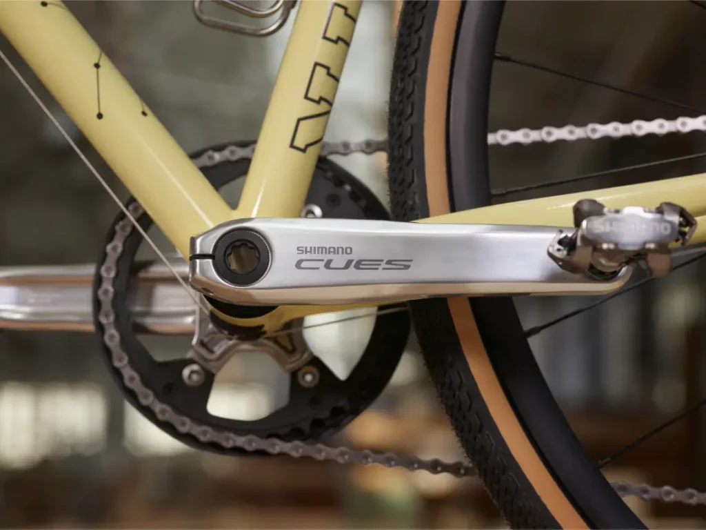 Primer plano de un plato y pedal Shimano CUES en una bicicleta amarilla, destacando la cadena, el plato y parte de la rueda trasera, con el acabado Aluminio Pulido que refleja la calidad vista en los grupos de carretera.