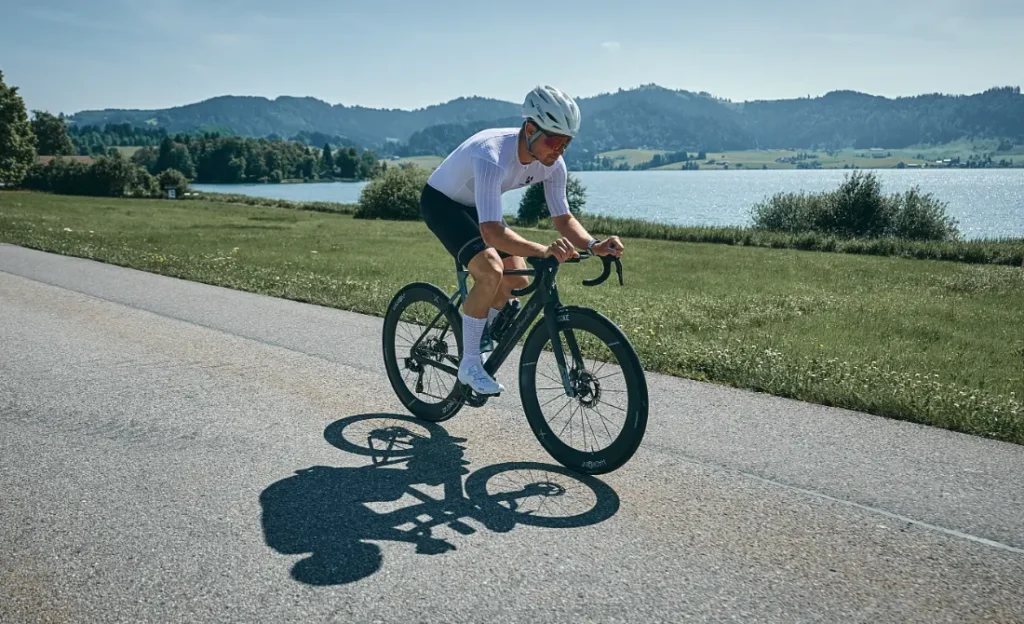 Un ciclista con casco y maillot blancos conduce una bicicleta de carretera HADRON³ Ultimate 650, conocida por su aerodinámica, por un camino asfaltado cerca de un lago, con colinas y vegetación de fondo.