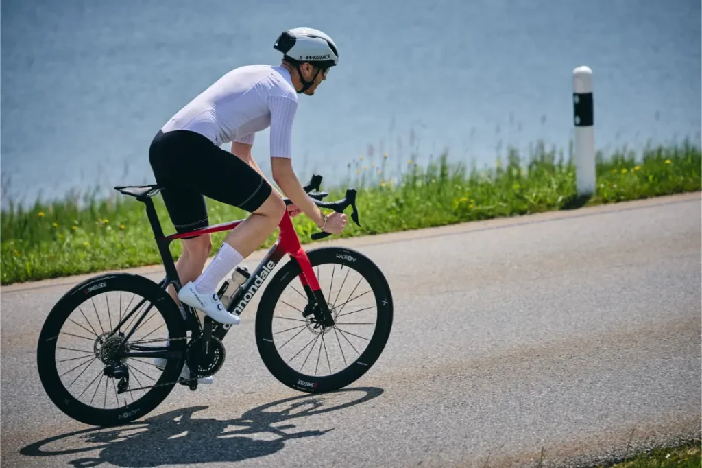 Un ciclista con maillot y casco blancos monta en una bicicleta de carretera roja y negra cerca del agua y la hierba, haciendo gala de aerodinámica con la Side suiza HADRON³ Ultimate 650 mientras se mantiene dentro del límite UCI.