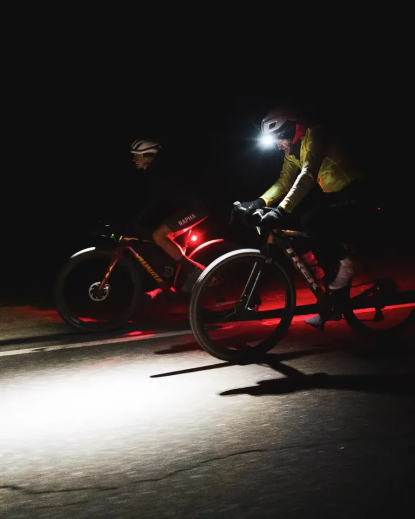 Dos ciclistas circulan de noche por una carretera oscura, iluminados por luces traseras rojas y un faro brillante, evocando el espíritu de aventura de States of Elevation o los legendarios viajes nocturnos de Kilian Jornet.