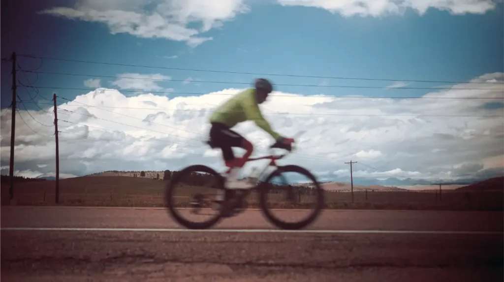 Un ciclista con casco y chaqueta verde recorre una carretera vacía bajo las líneas eléctricas y un cielo nublado, canalizando el espíritu de estados de elevación como Kilian Jornet.