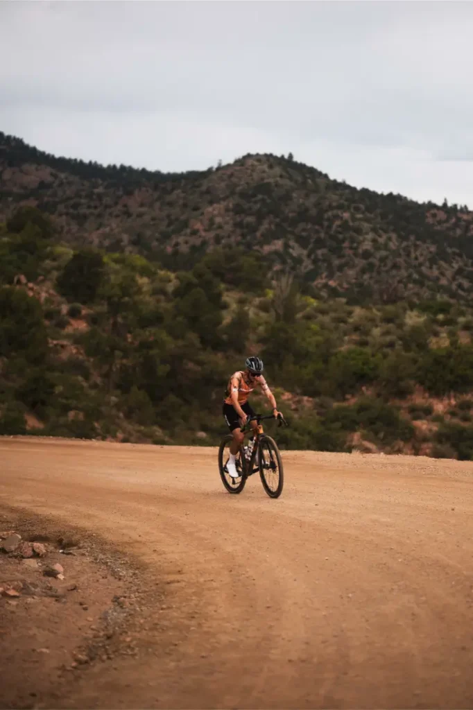 Un ciclista con casco conduce una bicicleta de gravilla por un sinuoso camino de tierra, canalizando el espíritu de States of Elevation - Kilian Jornet, con colinas y árboles de fondo bajo un cielo nublado.