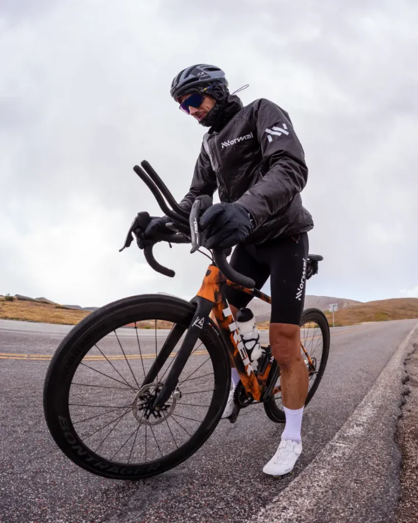 Un ciclista con casco, gafas de sol, chaqueta y guantes se para con una bicicleta de carretera en el arcén de una carretera vacía bajo un cielo nublado, evocando el espíritu de States of Elevation - Kilian Jornet.