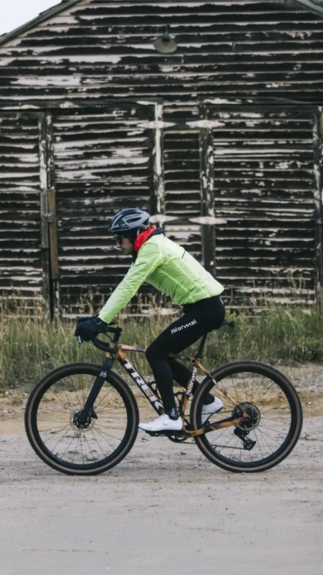 Una persona con casco y chaqueta brillante monta en bicicleta por un camino de tierra frente a un viejo edificio de madera desgastada, captando el espíritu de States of Elevation - Kilian Jornet.