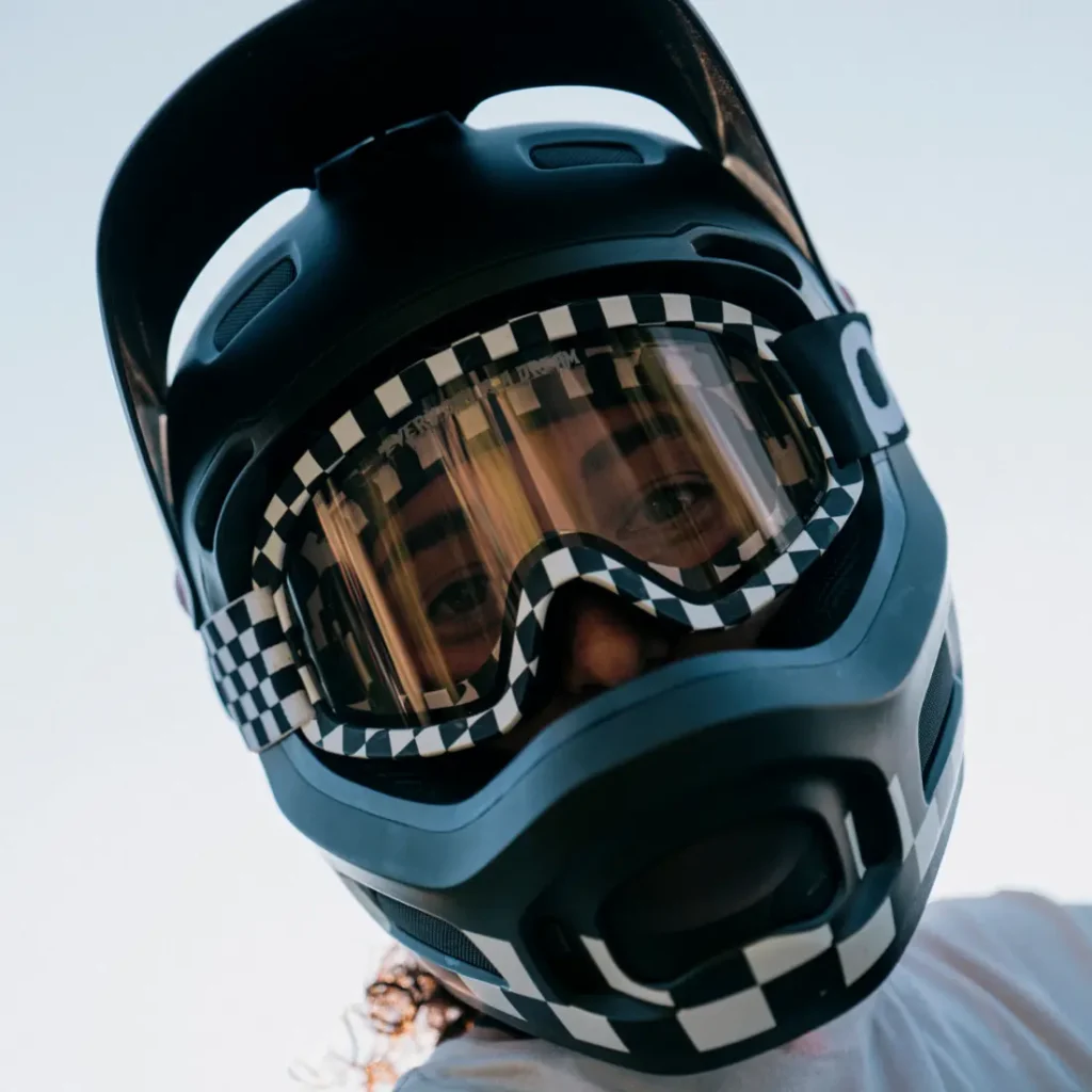Persona vestida con un casco Coron Air a cuadros blancos y negros y gafas Ora, fotografiada desde un ángulo bajo contra un cielo despejado.