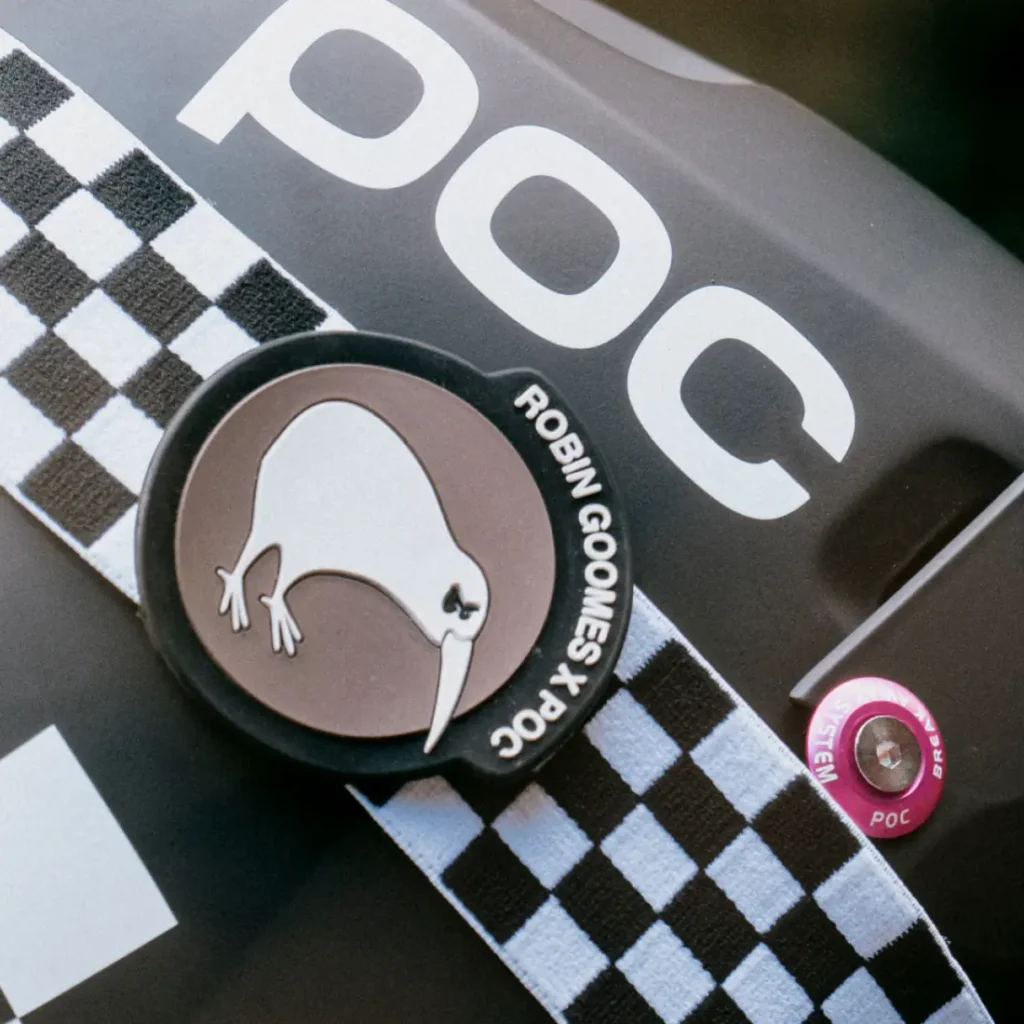 Primer plano de un casco Coron Air negro con la marca "poc", una correa a cuadros blancos y negros y una insignia de un pájaro kiwi con la inscripción "ROBIN GOOMES X POC".