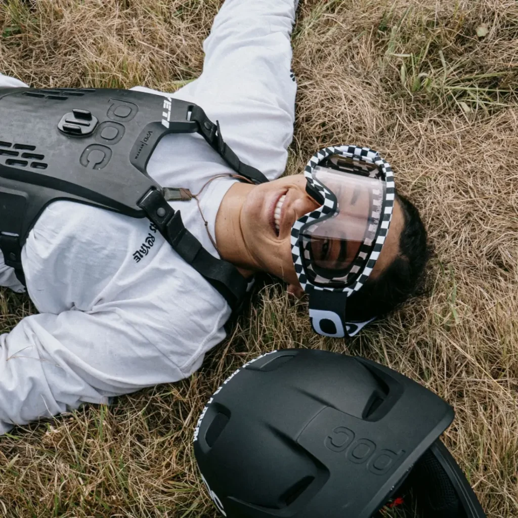 Persona con camisa blanca, equipo de protección y gafas Ora se tumba en la hierba seca junto a un casco negro Coron Air, sonriendo a la cámara, canalizando el espíritu aventurero de Robin Goomes.