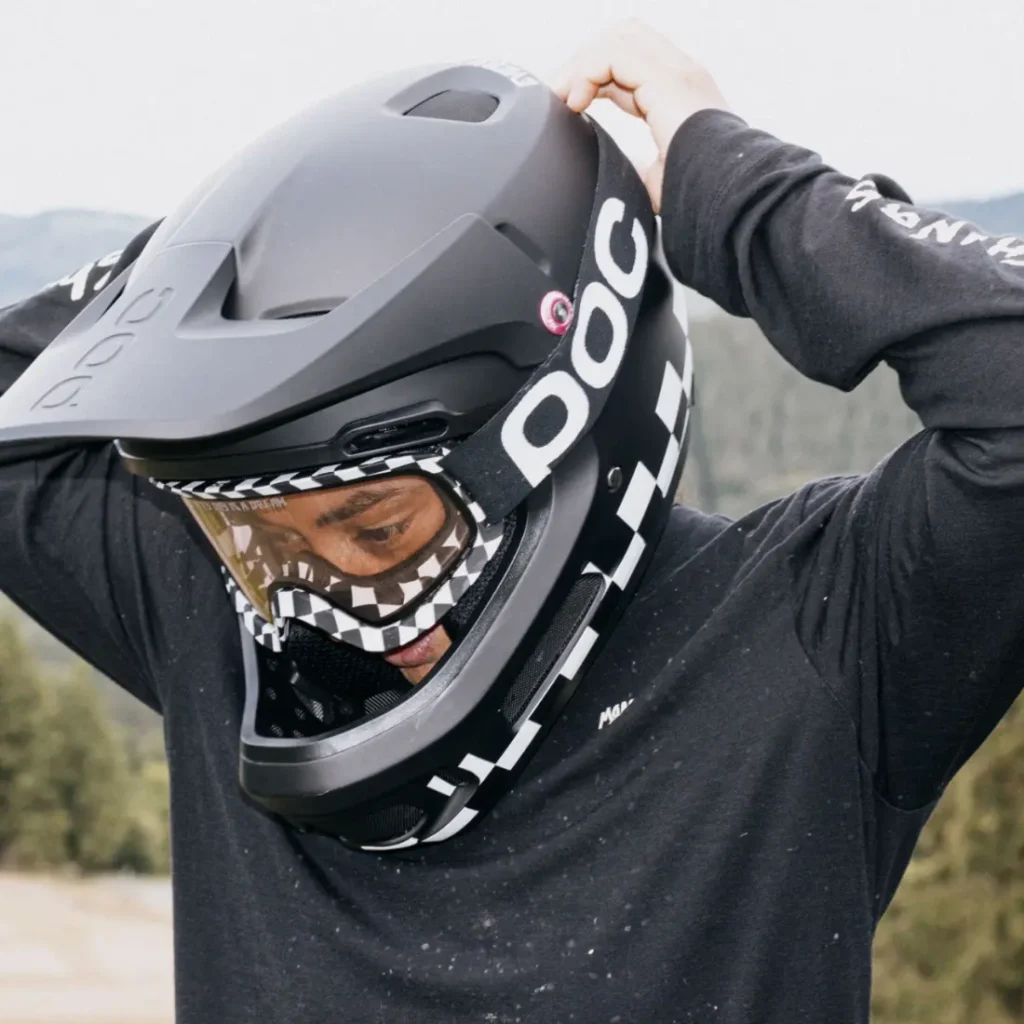 Una persona con camiseta negra de manga larga se ajusta un casco integral negro Coron Air POC con gafas Ora, al aire libre cerca de una zona boscosa.
