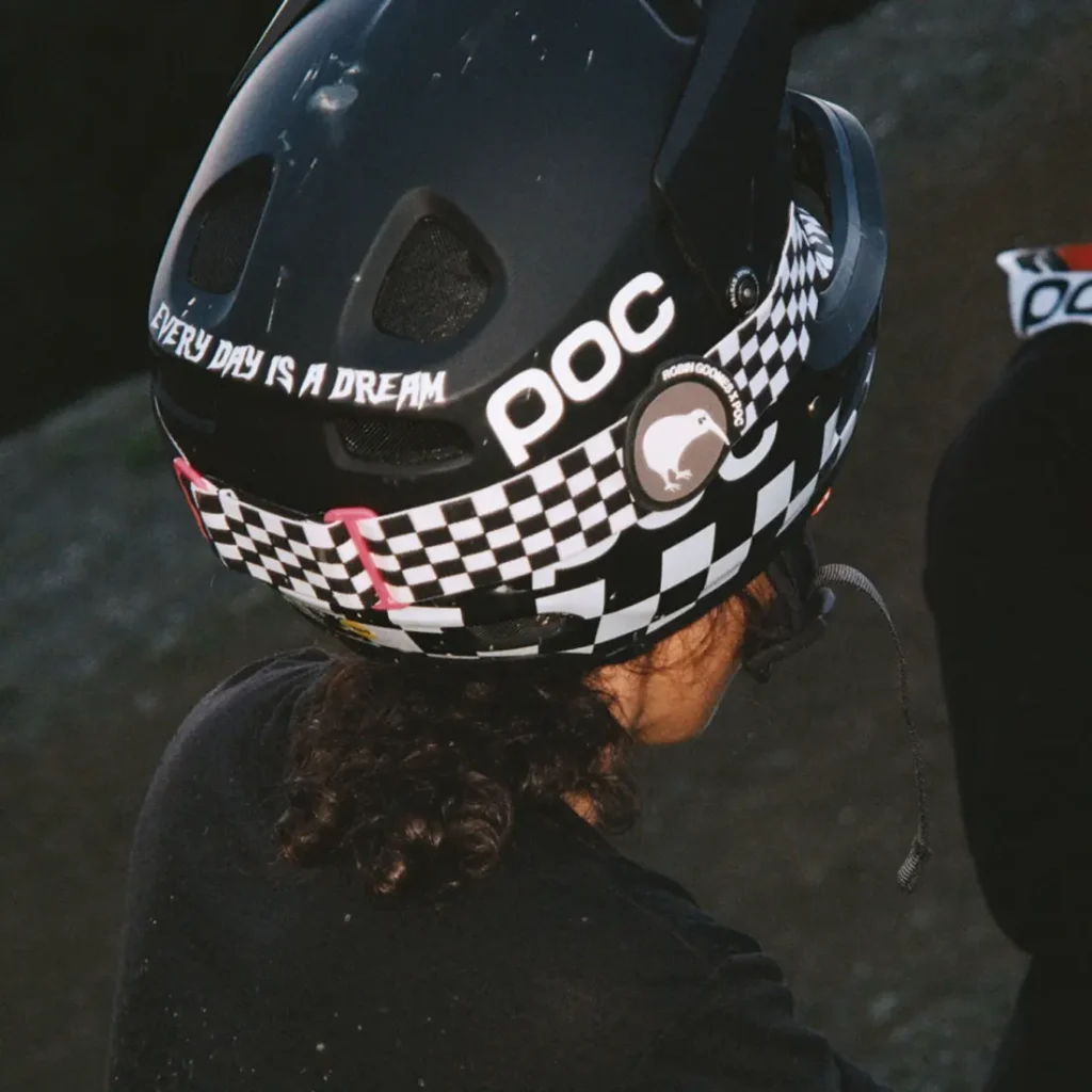Una persona lleva un casco negro de ciclista con un estampado de damero, el logotipo "POC" y el texto "EVERY DAY IS A DREAM" en la parte trasera, similar al icónico estilo de Robin Goomes y su casco favorito Coron Air.
