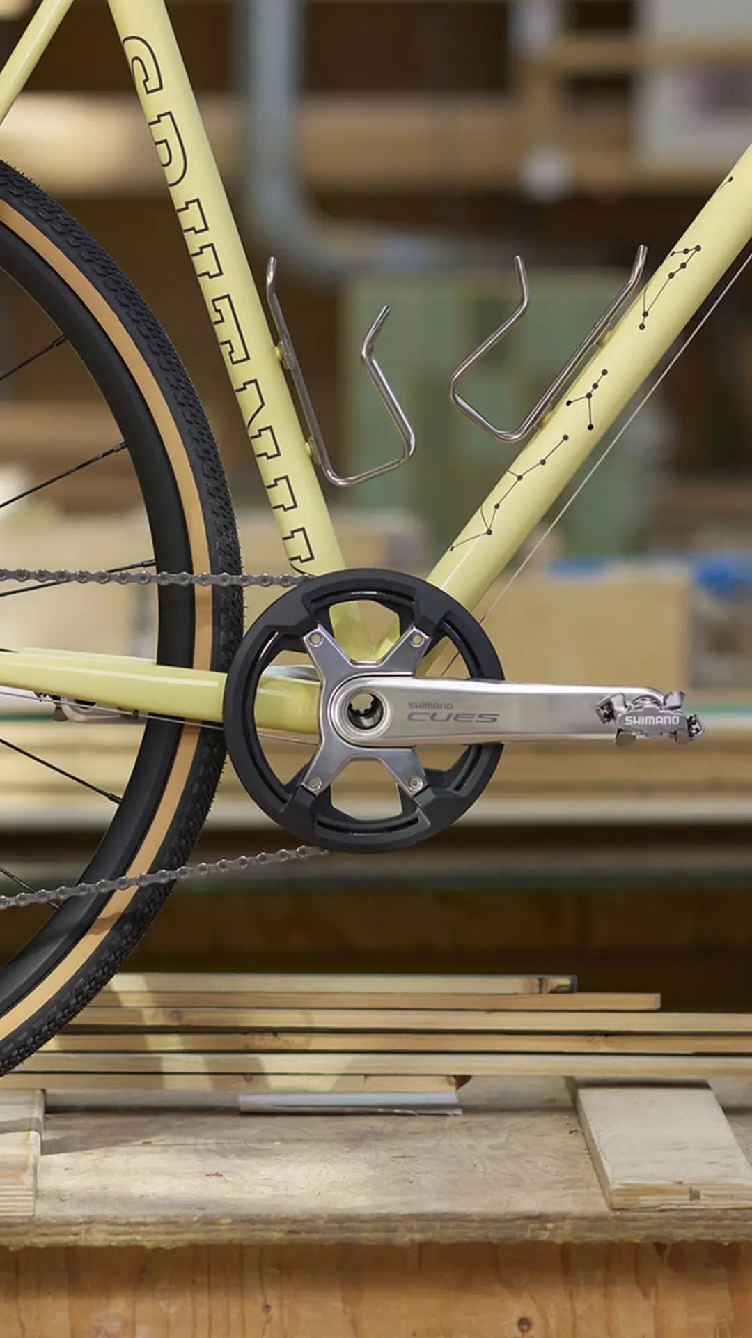 Primer plano del cuadro, la rueda trasera y los platos y bielas Shimano CUES de una bicicleta amarilla de aluminio pulido, con portabidones montados en el cuadro y tablones de madera al fondo.