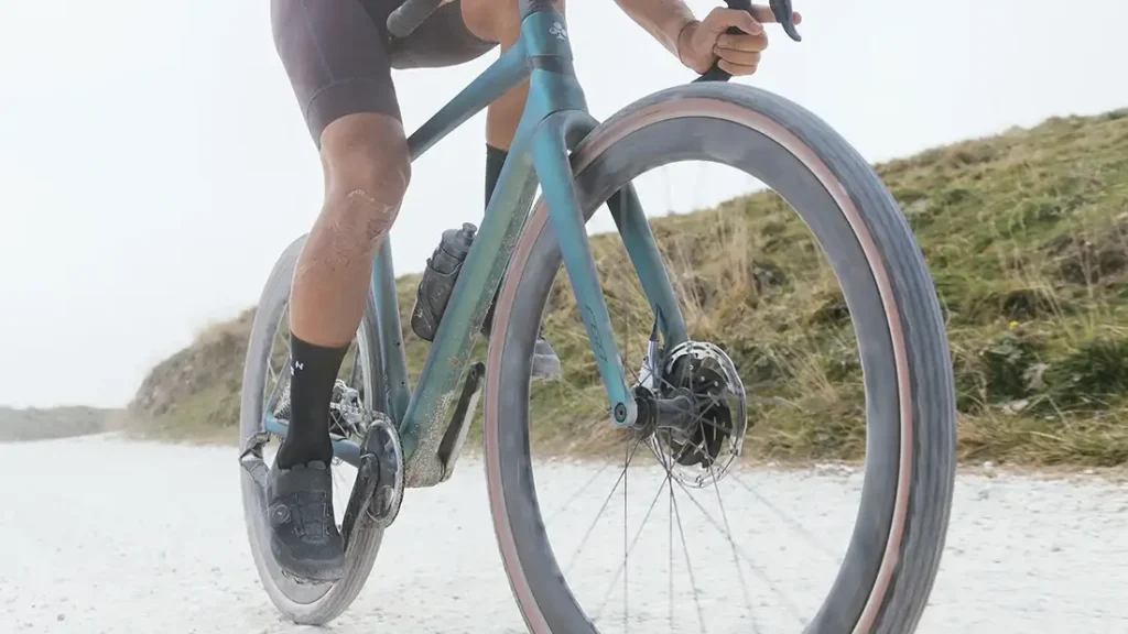 Primer plano de un ciclista montado en una bicicleta de grava azul equipada con neumáticos Pirelli Cinturato Gravel en una carretera polvorienta sin asfaltar con terreno herboso al fondo.