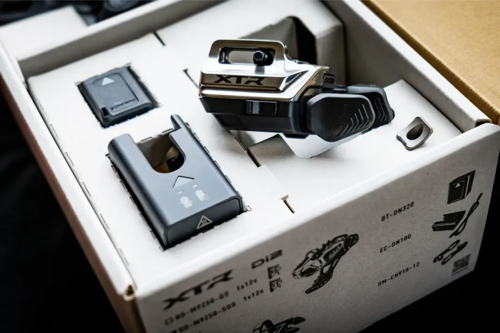 Un juego de componentes para bicicleta Shimano XTR, que forma parte de los kits de actualización Shimano premium, dentro de una caja de cartón, con un cambio electrónico, una batería y accesorios perfectamente organizados en un embalaje ajustado.