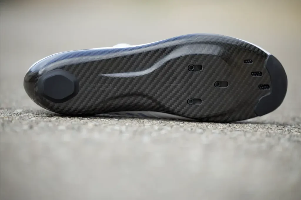 Vista en primer plano de la suela de fibra de carbono de las Zapatillas Q36.5 para ciclismo con orificios de fijación para calas, diseñadas para obtener el máximo rendimiento, apoyada sobre una superficie rugosa al aire libre.