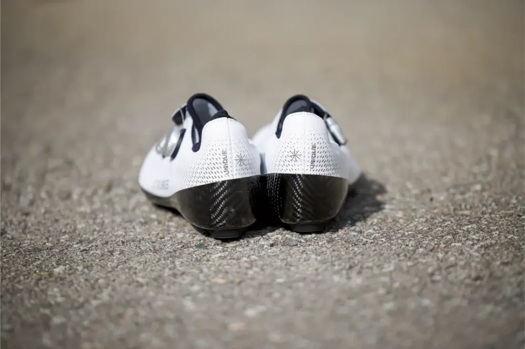 Un par de Zapatillas Q36.5 Unique Pro blancas con suela negra se colocan una al lado de la otra sobre una superficie pavimentada, vistas desde atrás, mostrando su diseño orientado al rendimiento.