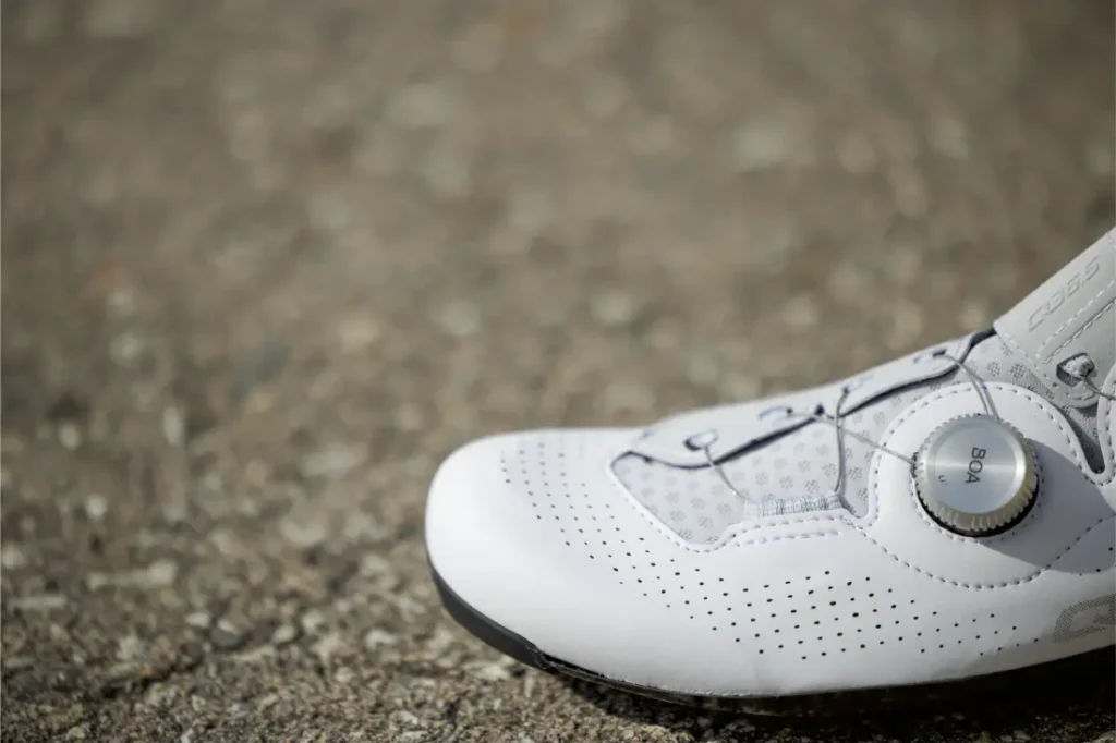 Primer plano de unas Zapatillas Q36.5 Unique Pro blancas de ciclismo con sistema de cierre de dial BOA sobre una superficie de asfalto, diseñadas para obtener el máximo rendimiento.