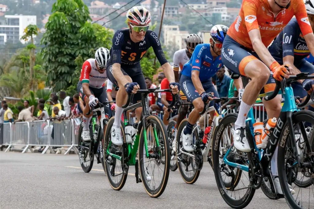 Ciclistas con equipamiento profesional, entre ellos el grimpeur-cróner Abel Balderstone, compiten en una carrera de carretera mientras los espectadores observan detrás de unas barricadas en una calle de la ciudad.
