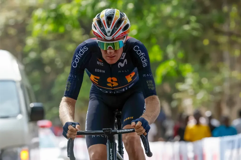 Un ciclista, como Abel Balderstone, con casco, gafas de sol y uniforme oscuro de equipo monta en bicicleta durante una carrera, con espectadores y vegetación borrosos al fondo.