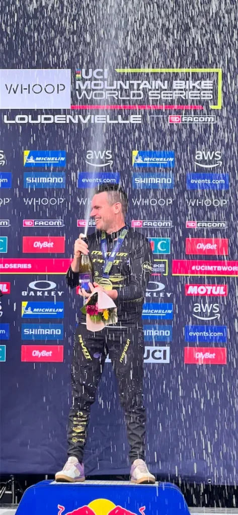 Un piloto de carreras, Jordi Bagó, en traje de carreras, de pie en un podio con una botella en la mano y un micrófono, mientras salpica champán, dando una entrevista Jordi Bagó con logotipos de patrocinadores en el telón de fondo detrás de él.