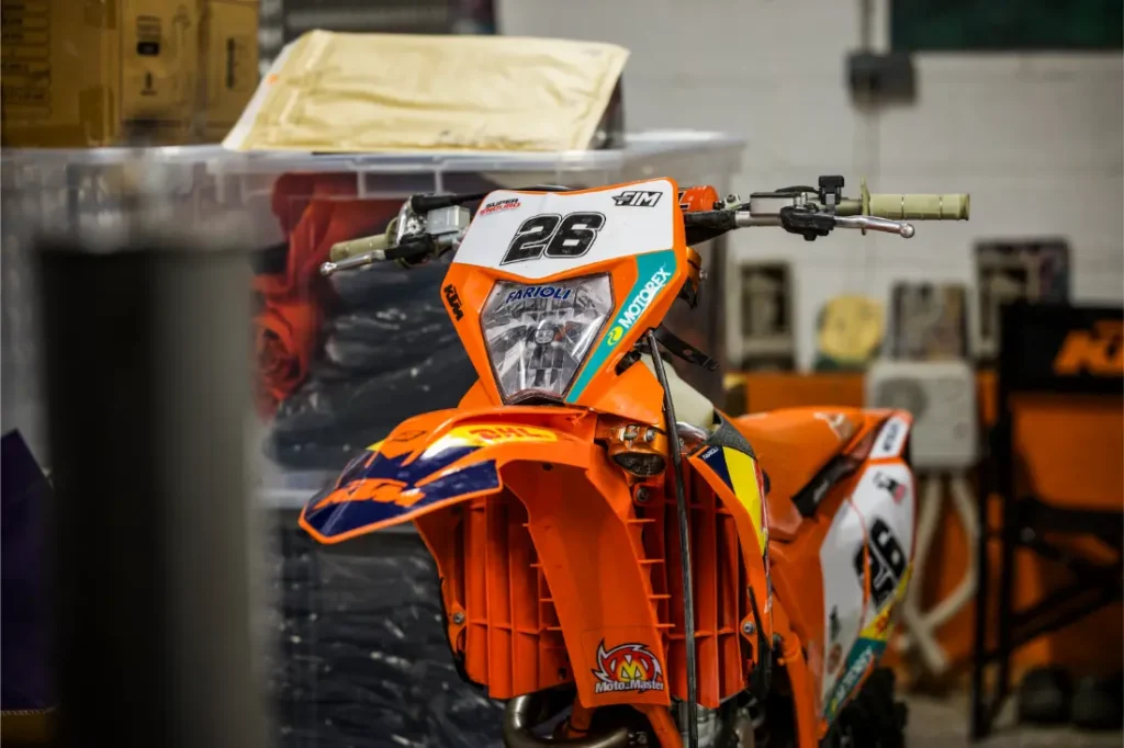 Una moto de cross naranja con el número 26 en la parte delantera está aparcada en el interior cerca de estanterías y cajas de almacenaje, similares a las que se ven en una Entrevista a Josep García.