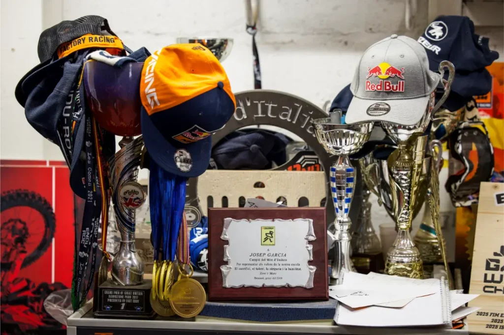Una exhibición de trofeos, medallas, placas y gorras -incluida una gorra de Red Bull- dispuesta en una estantería con diversos recuerdos y artículos de carreras de la Entrevista a Josep García.