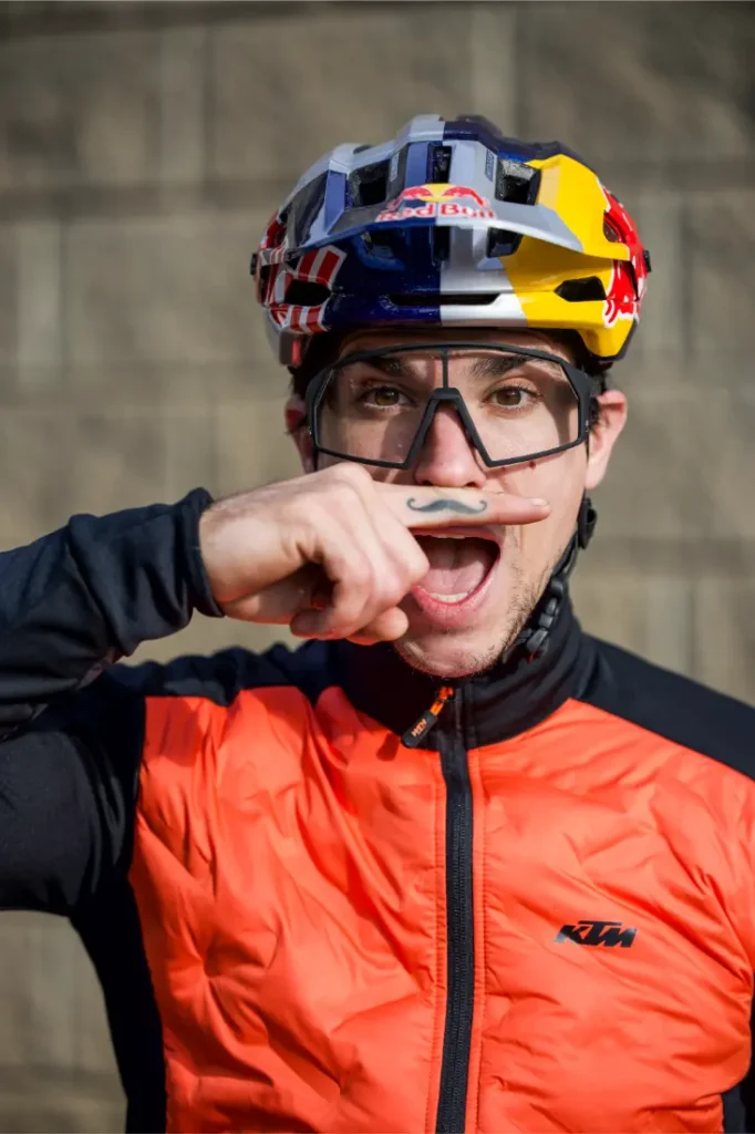 Un hombre con casco de moto y gafas sostiene un objeto en forma de bigote bajo la nariz, con la boca abierta, vestido con una chaqueta naranja de KTM durante una entrevista a Josep García.