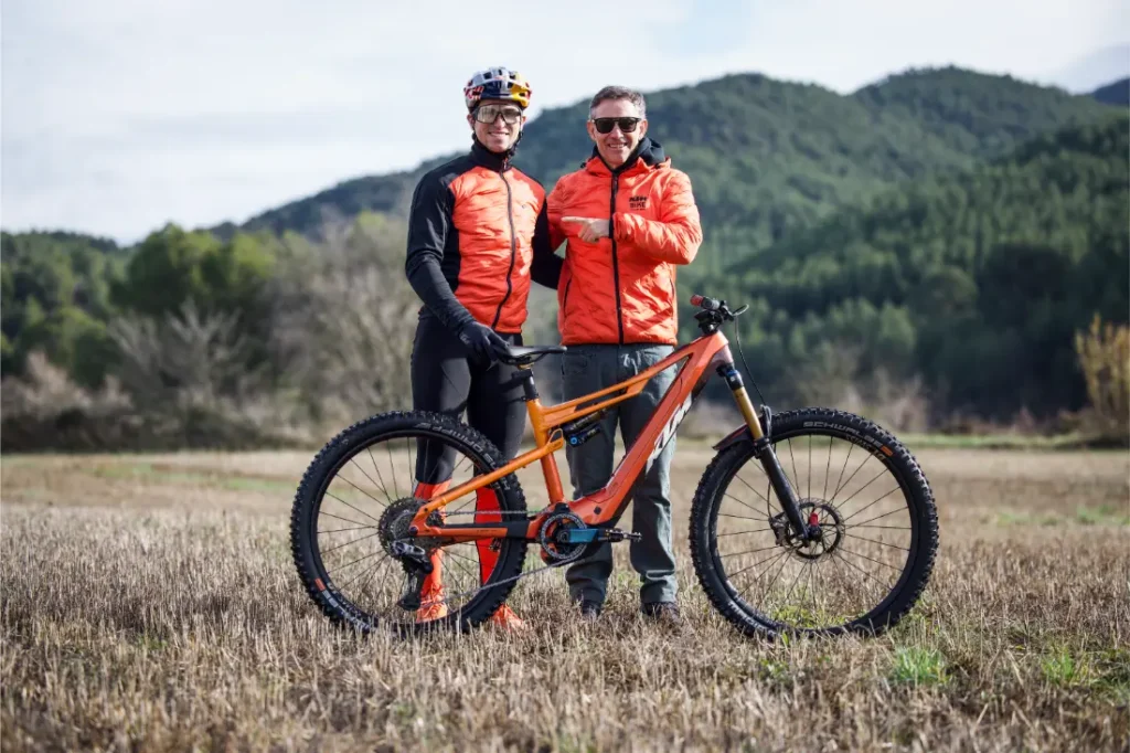 Dos hombres con chaquetas naranjas se paran al aire libre en un campo con una bicicleta de montaña, con árboles y colinas detrás de ellos durante una entrevista a Josep García.