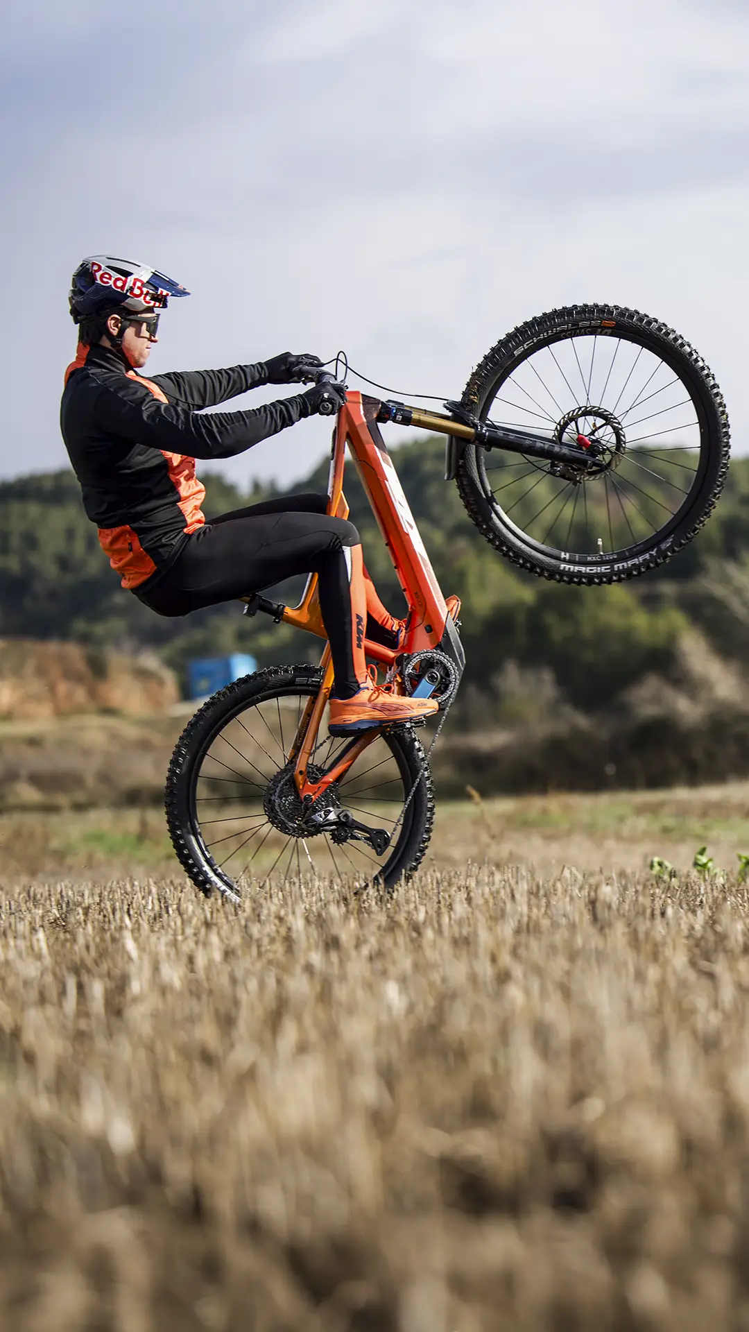 Una persona con casco realiza un caballito en una bicicleta de montaña naranja en un campo abierto con árboles al fondo.