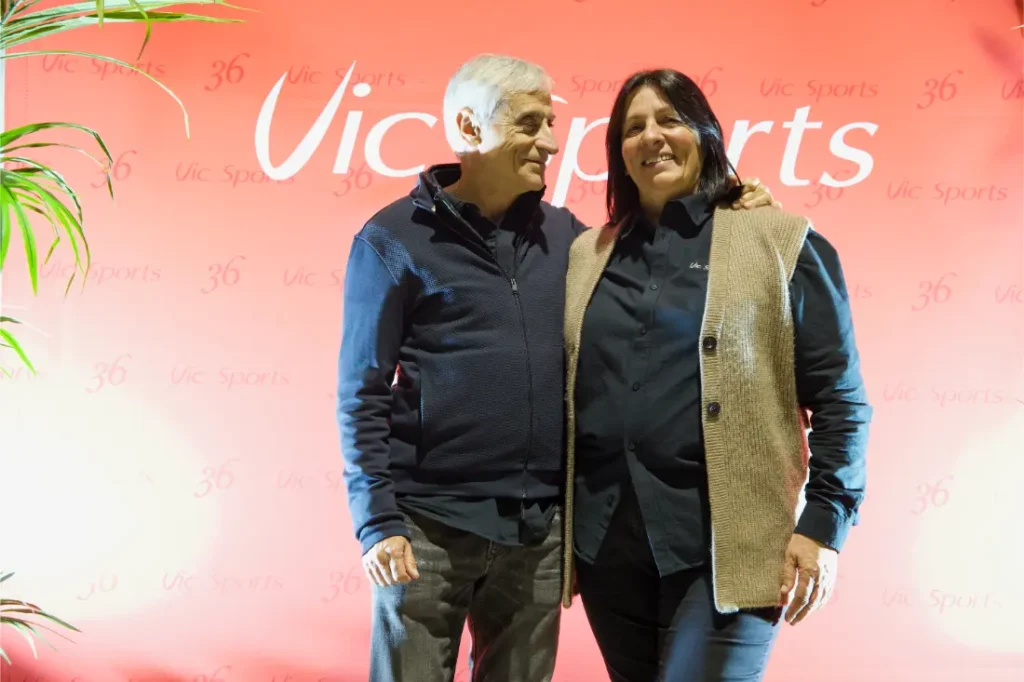 Dos personas permanecen juntas, sonrientes, delante de un atrevido telón de fondo rojo de Vic Sports, mostrando su entusiasmo por el acontecimiento Vic Sports.