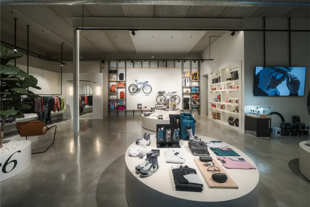 Interior de tienda moderna con mesas redondas de exposición de ropa y accesorios de Vic Sports, estanterías con bolsos y una bicicleta en la pared del fondo, y una pantalla que muestra un anuncio.