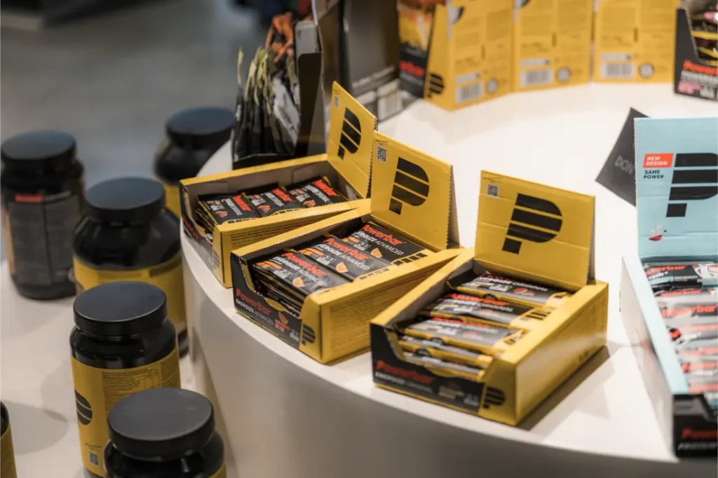 Cajas de barritas de proteínas PowerBar se exhiben en un mostrador blanco curvado junto a varios contenedores negros de suplementos, mostrando las mejores opciones de nutrición en Vic Sports.