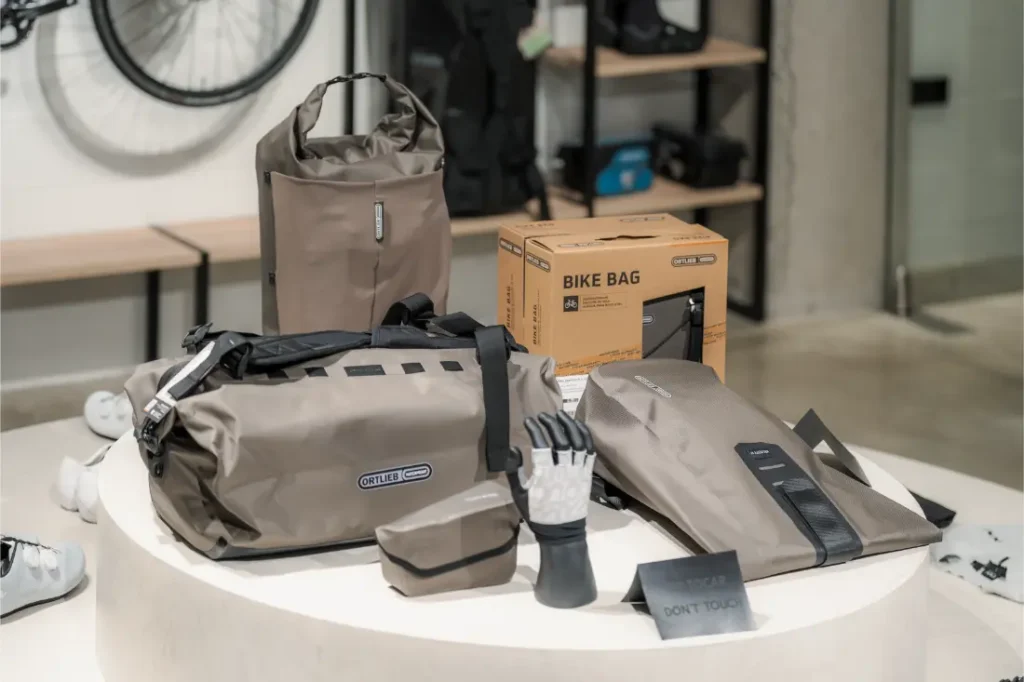 Un expositor de bolsas y accesorios Ortlieb de color caqui, que incluye una bolsa de viaje, un bolso tote, guantes y una bolsa para bicicletas en caja, dispuestos sobre una mesa redonda blanca en la tienda de Vic Sports.