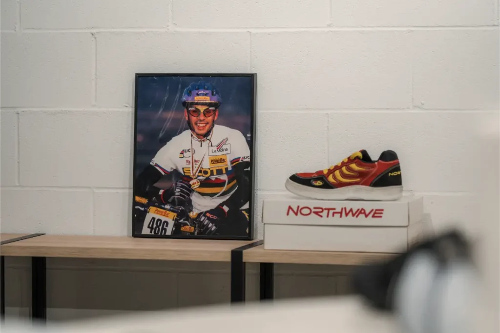 Una foto enmarcada de un ciclista y unas zapatillas Northwave rojas y amarillas descansan sobre dos cajas apiladas de Northwave de Vic Sports, expuestas en una estantería de madera contra una pared blanca.