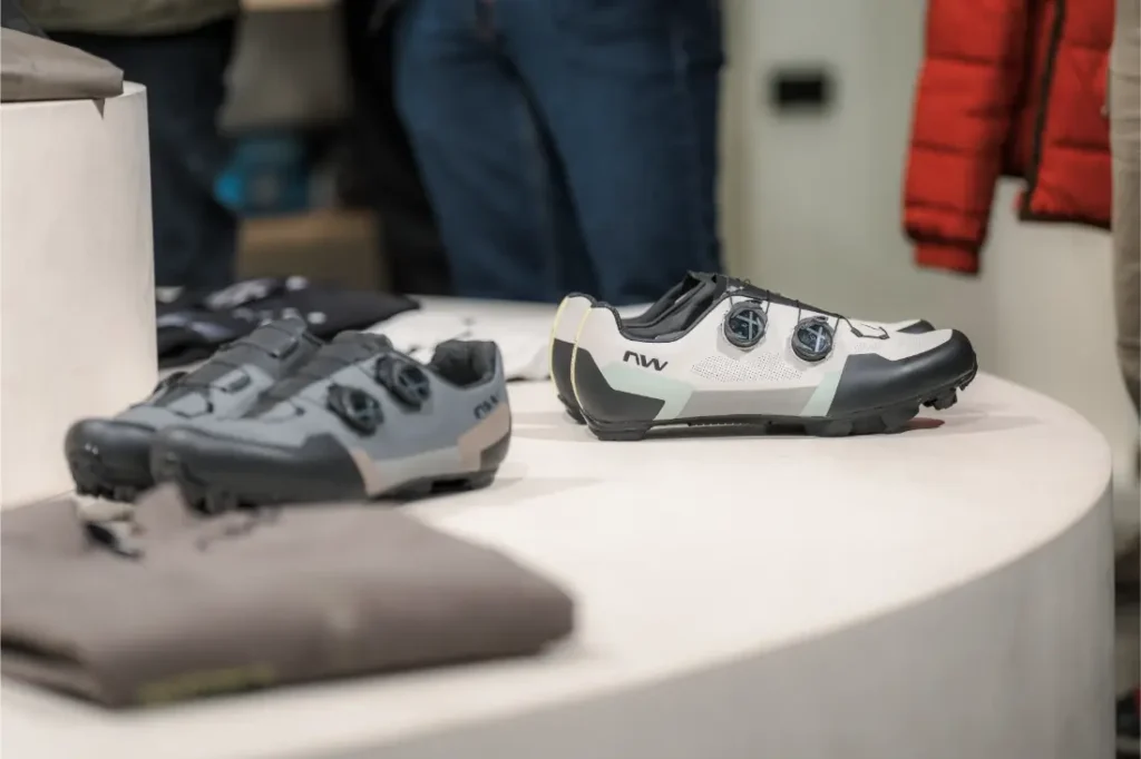 Dos zapatillas de ciclismo Vic Sports con cierre de esfera se exponen sobre una superficie blanca, con prendas de vestir dobladas visibles en las proximidades.