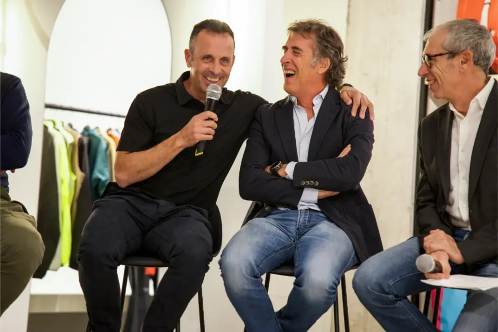 Tres hombres sentados en taburetes durante una mesa redonda de Vic Sports; uno de ellos sostiene un micrófono y sonríe, mientras los demás escuchan y ríen.