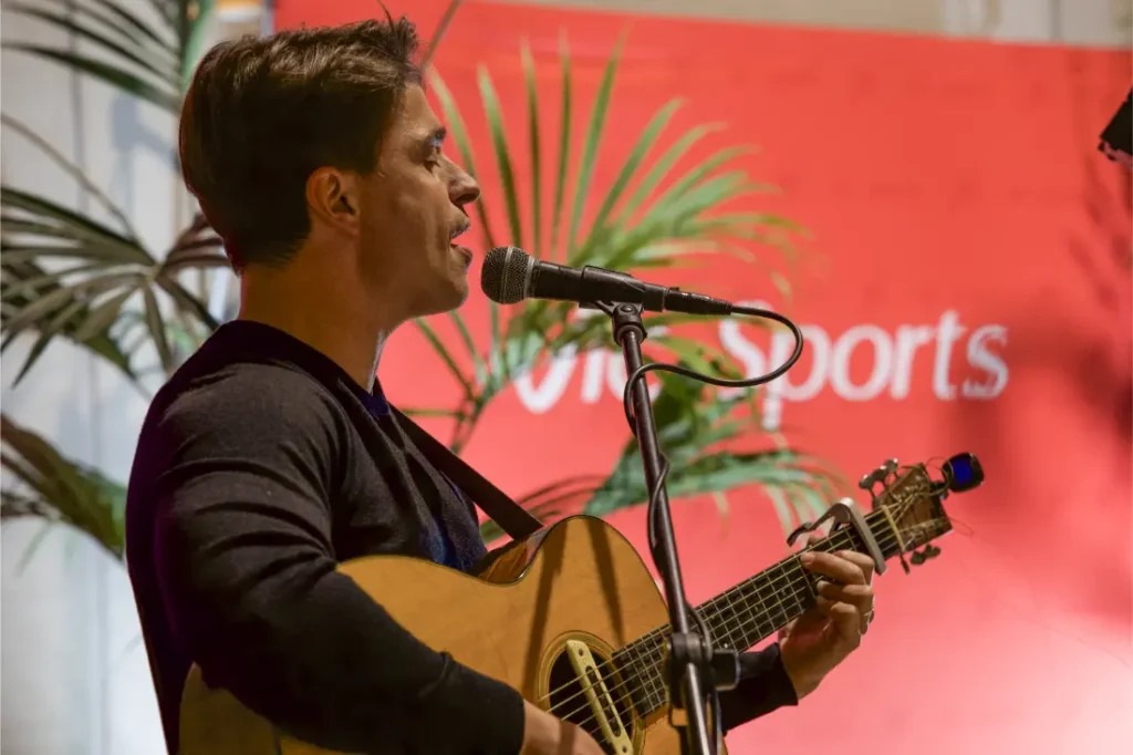 Un hombre toca una guitarra acústica y canta en un micrófono delante de un telón de fondo rojo con "Vic Sports" y "Viaplay Sports", rodeado de macetas verdes.