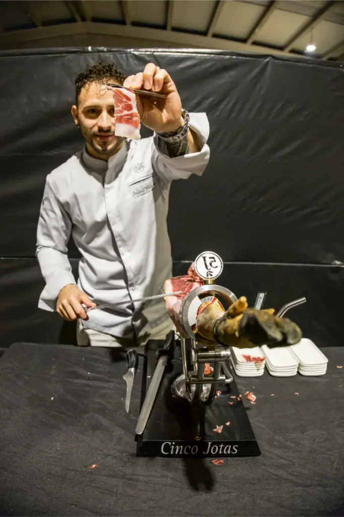 Un cocinero con bata blanca sostiene una loncha de jamón curado, de pie detrás de una pata de jamón en un soporte de corte con la etiqueta "Cinco Jotas", que recuerda la destreza y precisión de las competiciones de Vic Sports.