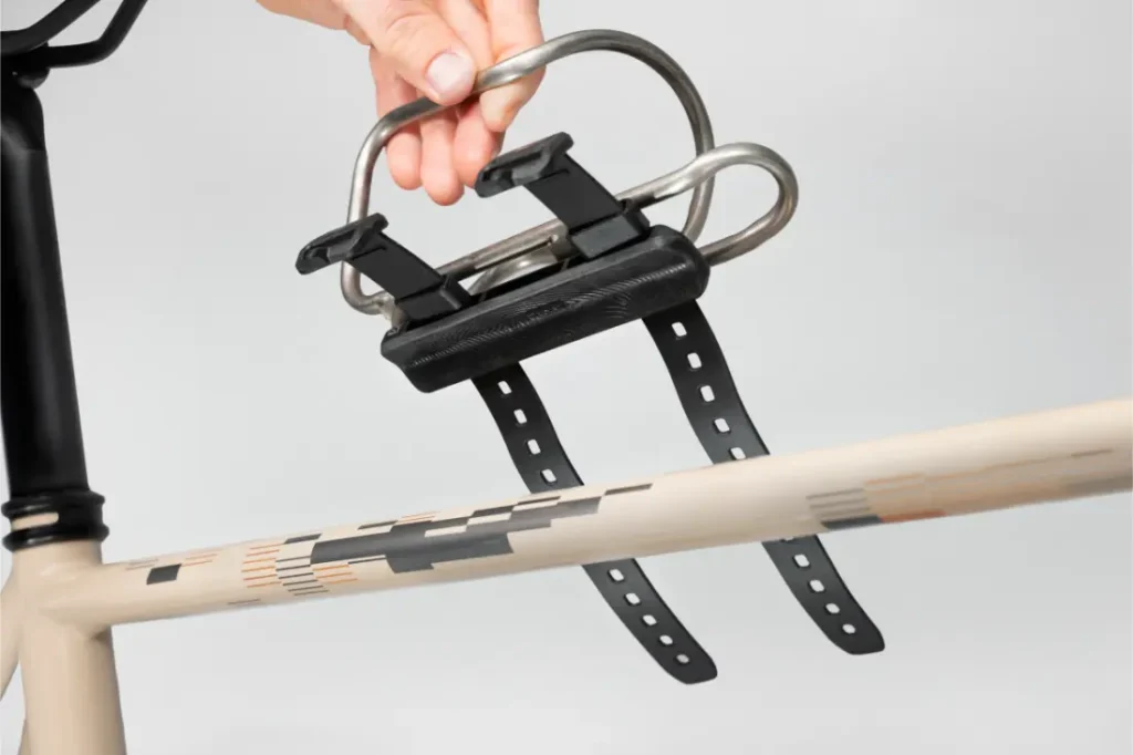 Una mano instala un portabidón metálico Tailfin Hydromount con dos correas en el tubo superior del cuadro de una bicicleta beige.