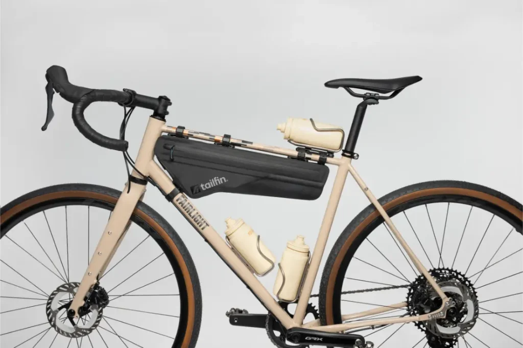 Una bicicleta de grava beige con manillar negro, una bolsa de cuadro y tres bidones de agua beige montados en el cuadro con soportes hidráulicos Tailfin, sobre un fondo liso.