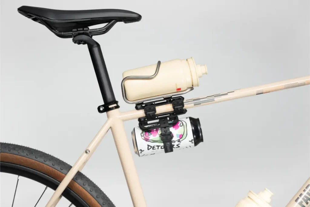 Un cuadro de bicicleta de color beige con dos bidones de agua fijados al tubo del sillín mediante Tailfin Hydromounts y soportes de plástico, colocados por encima de la rueda trasera.