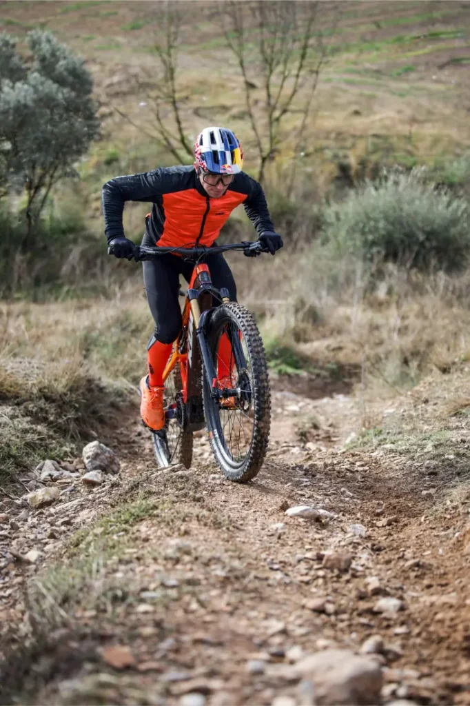 Un ciclista vestido de naranja y negro, inspirado en Entrevista a Josep García, conduce una bicicleta de montaña cuesta arriba por un sendero de tierra rocosa en un entorno rural al aire libre.