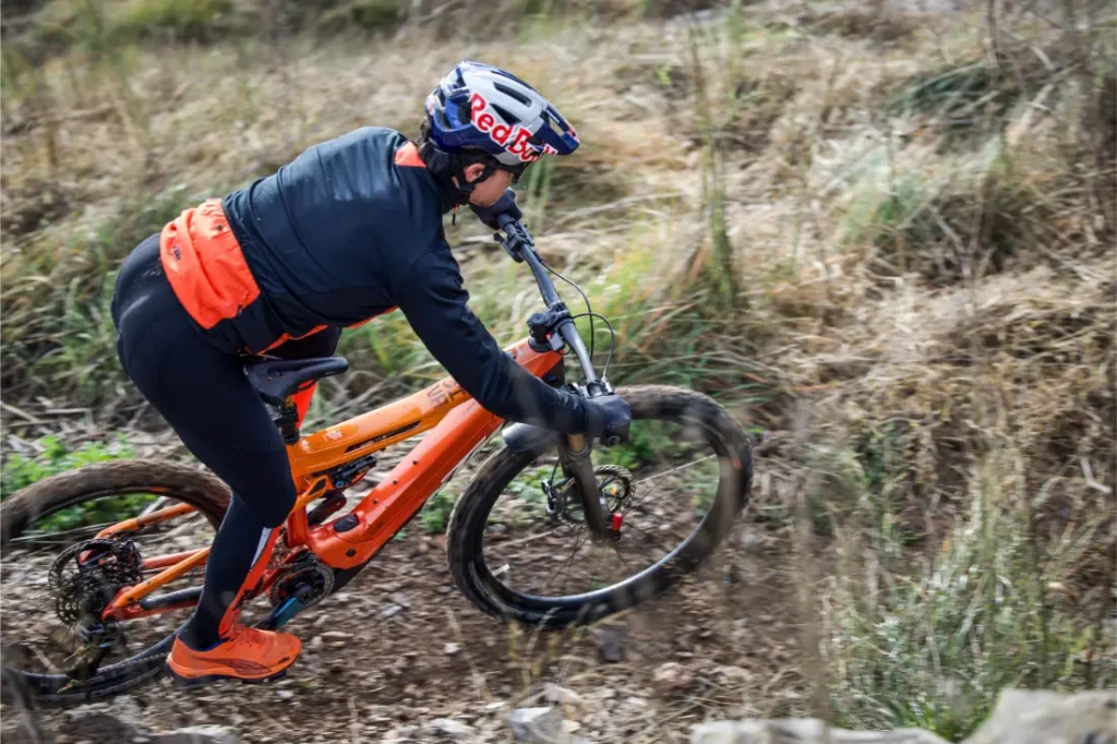 Un ciclista con casco y ropa naranja conduce una bicicleta de montaña por un sendero de tierra a través de un terreno cubierto de hierba, que recuerda escenas de la Entrevista a Josep García.