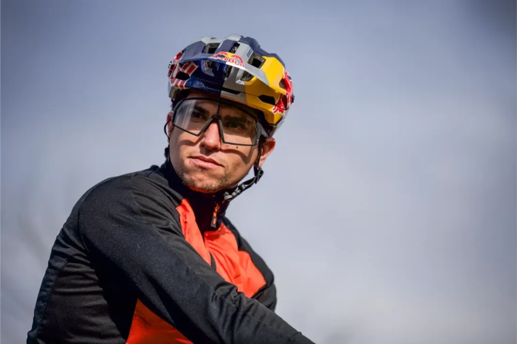 Un ciclista con un casco de colores, gafas protectoras y una chaqueta roja y negra mira a un lado sobre un fondo exterior difuminado, como si se preparara para una Entrevista a Josep García.