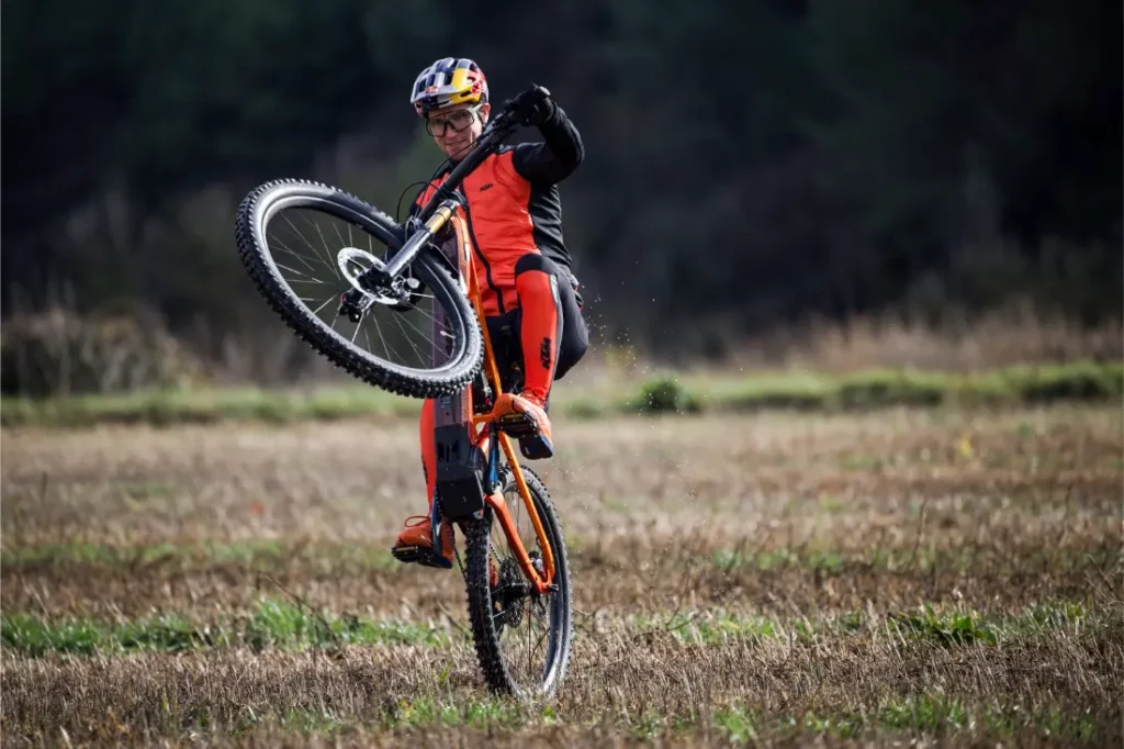 Persona con indumentaria ciclista naranja haciendo un caballito en una bicicleta de montaña en un campo abierto cubierto de hierba, con árboles difuminados al fondo, como en las escenas de una entrevista a Josep García.