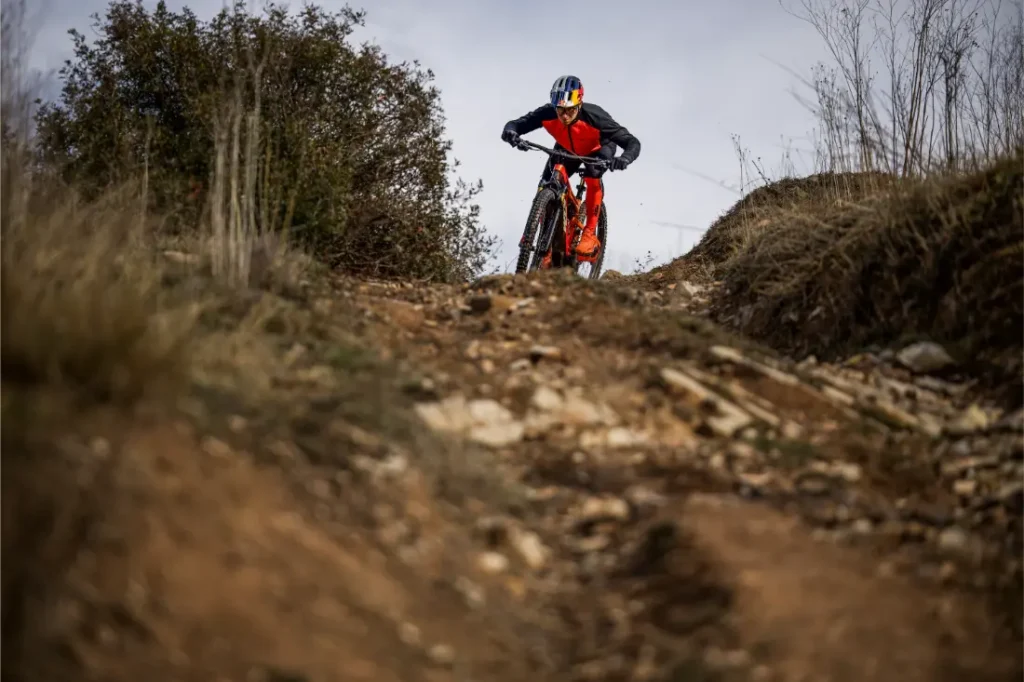 Un ciclista de montaña vestido de rojo y negro desciende por un sendero pedregoso de tierra rodeado de hierba seca y arbustos, que recuerda el accidentado terreno comentado en la entrevista a Josep García.