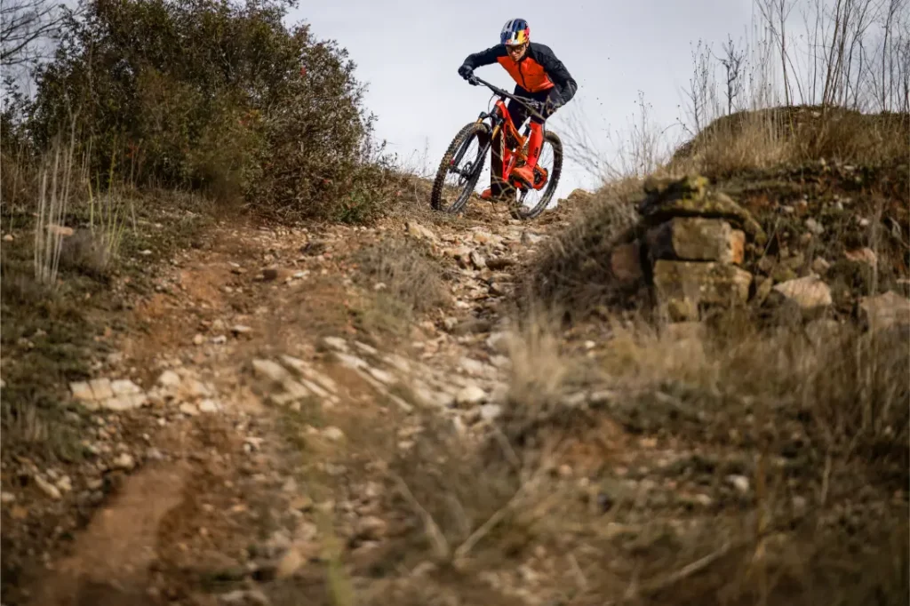 Una persona con ropa de ciclista baja en bicicleta de montaña por un sendero de tierra rocosa rodeado de hierba seca y arbustos, que recuerda a las escenas de una Entrevista a Josep García.